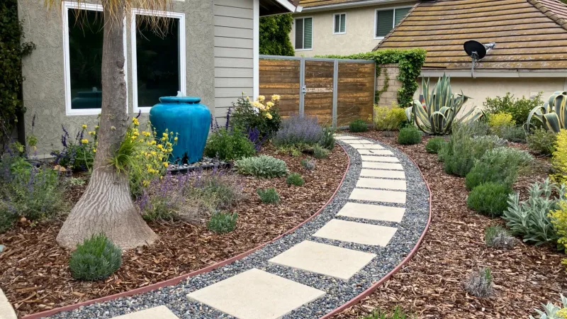Curved garden path with stepping stones surrounded by mulch, plants, and a blue planter adds a splash of color.