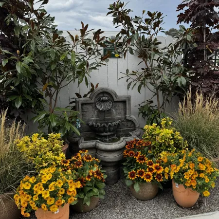 Stone garden fountain surrounded by vibrant yellow and orange flowers in pots, against a white fence under cloudy sky.