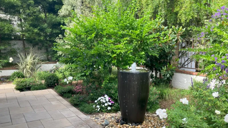 Garden scene with a black cylindrical water fountain surrounded by green plants and white flowers on a stone bed.
