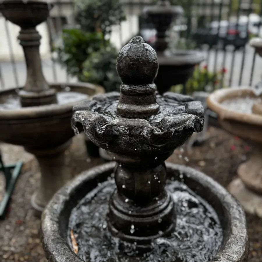 Close-up of a stone garden fountain with water flowing, blurred background of plants and other fountains.