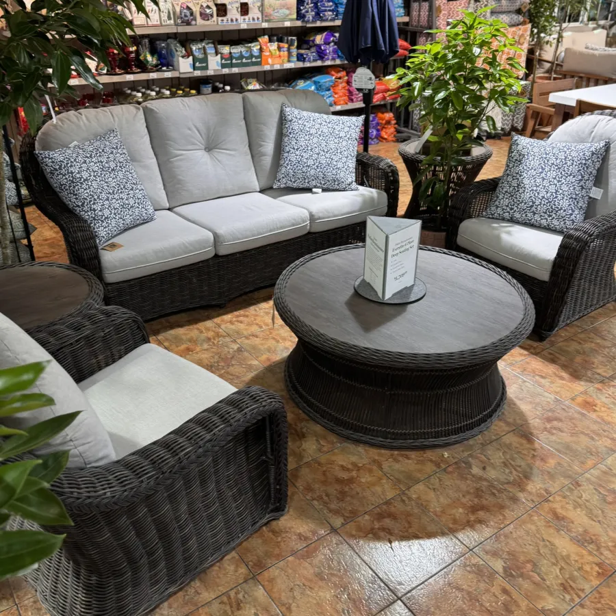 Wicker patio furniture set with light cushions and patterned pillows around a round coffee table on tiled floor indoors.