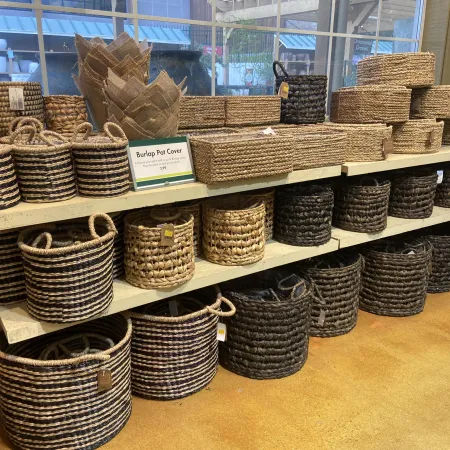 Display of various woven baskets and burlap pot covers arranged on wooden shelves in a store.