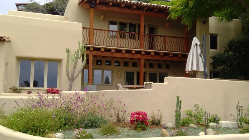 Southwestern style two-story house with beige stucco walls, wooden balcony, and desert landscaping with flowers and cacti.