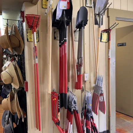 Display of various gardening tools including rakes, shovels, pruners, and hats hanging on a store wall.