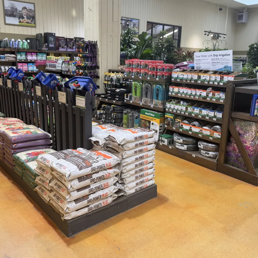 Garden supply store aisle with bags of soil, irrigation tools, hoses, and gardening products neatly displayed on shelves.