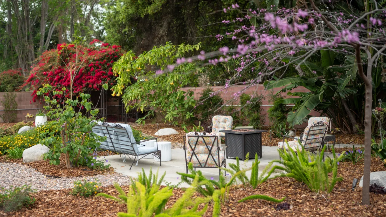 Outdoor patio seating area surrounded by lush garden plants, colorful flowers, and mulch-covered paths