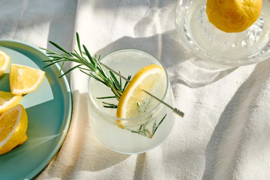 Refreshing lemon drink with rosemary sprig served on a light cloth with lemon wedges and a juicer.