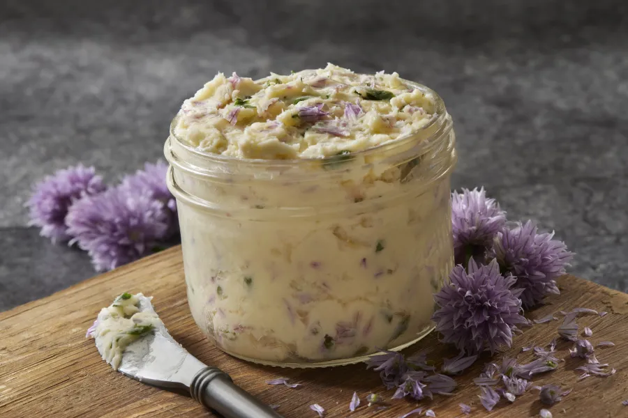 Jar of herb butter with purple chive flowers on wooden board and gray background.