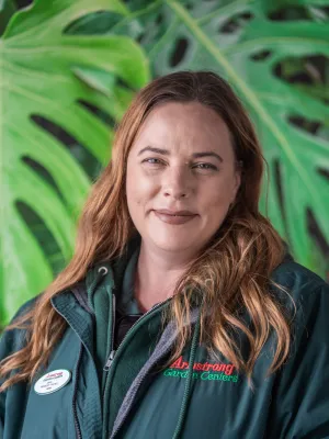 Smiling woman with long hair wearing Armstrong Garden Centers jacket standing in front of large green leaves.