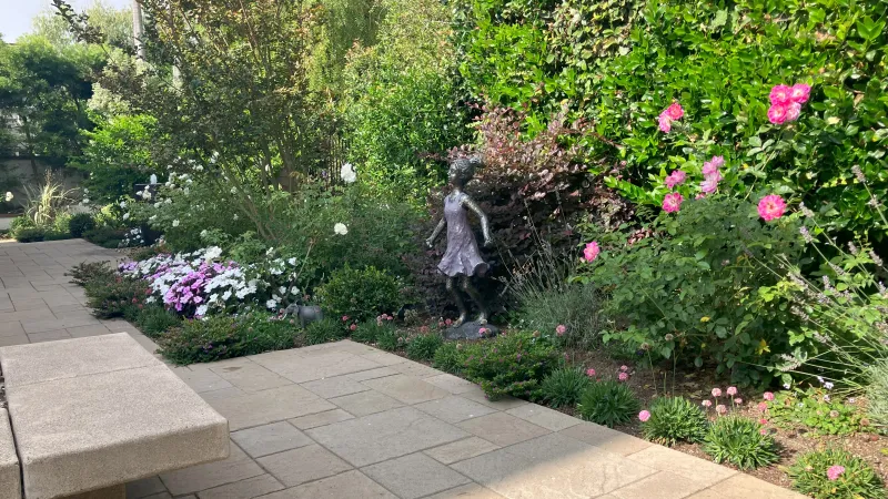 Sunny garden with stone bench, colorful flowers, lush greenery, and a bronze statue of a girl in a dress.